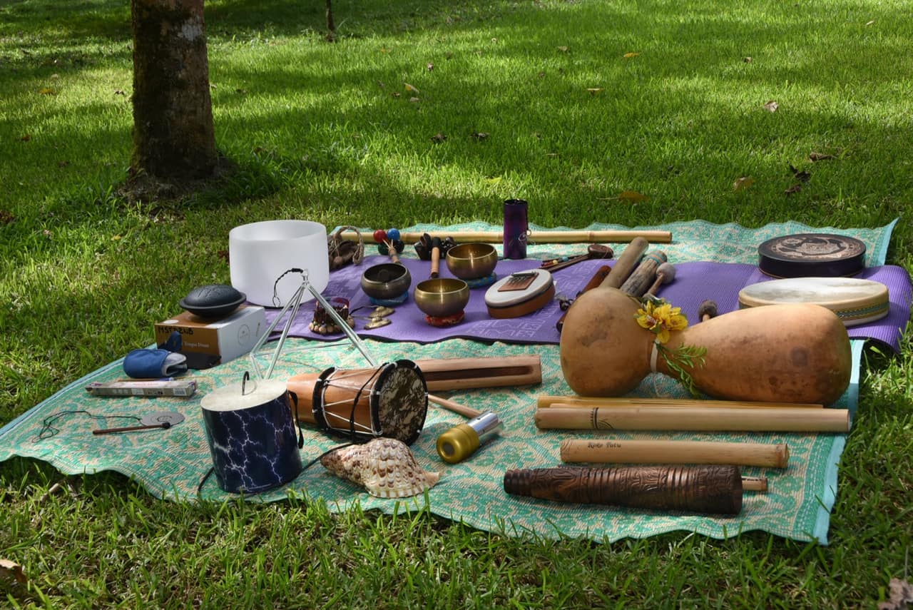 La collection complète des instruments KokoPatu disposée sur des nattes tressées : bol de cristal, bols tibétains, tongue drum, tambours, calebasses, flûtes de bambou et conques